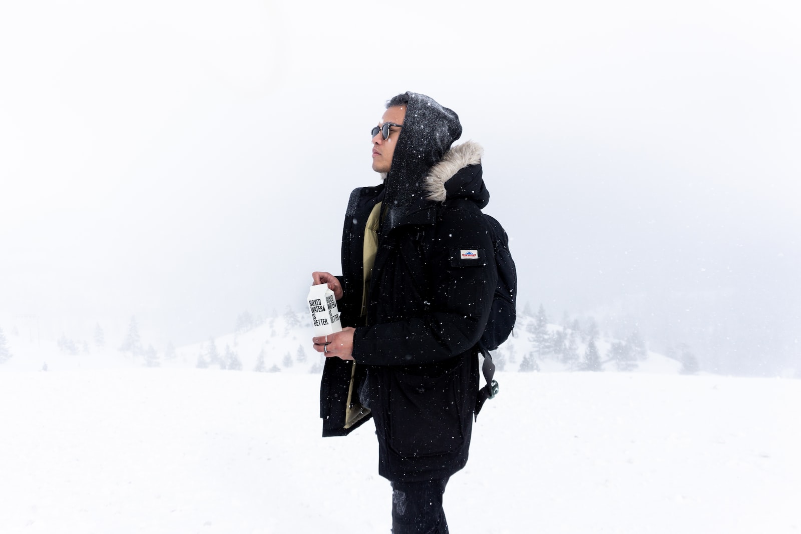 person wearing black hooded jacket holding boxed water while standing on snowy field during daytime